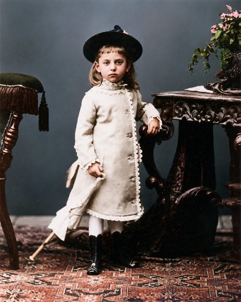 Childhood photograph of Rainer Maria Rilke with long hair, wearing a hat and a light-colored dress, standing beside an ornate table.